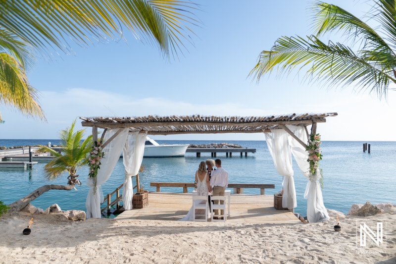 Couple stands together on a dock at Mood Beach in Curacao during their wedding ceremony under clear skies and near calm waters