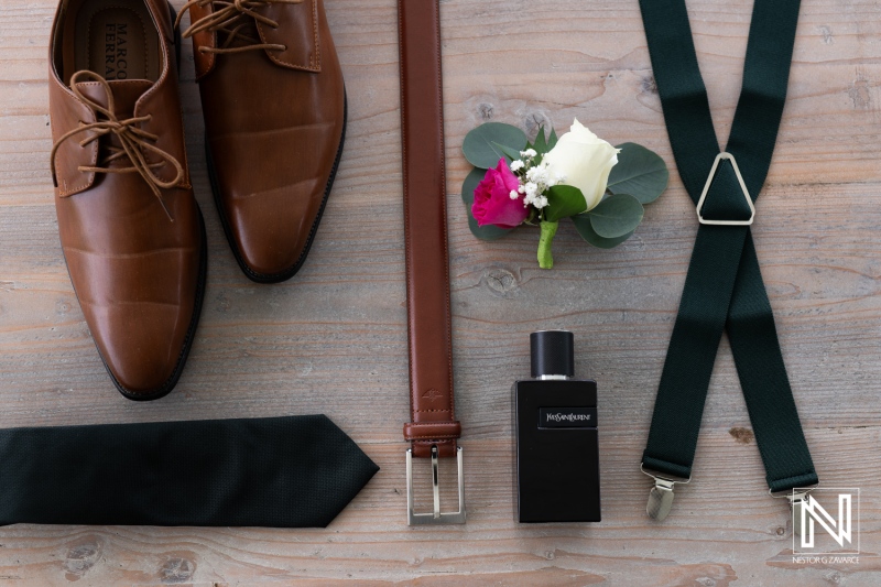 Elegant Groom\'s Attire Laid out on a Wooden Surface, Featuring Shoes, Belt, Tie, Suspenders, Fragrance, and a Boutonnière, Prepared for a Wedding Ceremony