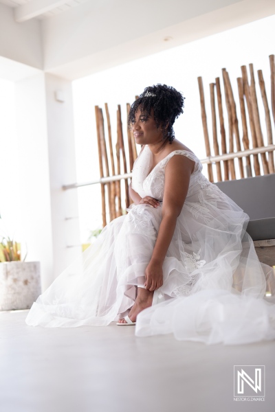 A Bride in a Flowing White Gown Adjusts Her Shoe While Seated Indoors, Surrounded by Natural Light and Wooden Accents in a Modern Setting