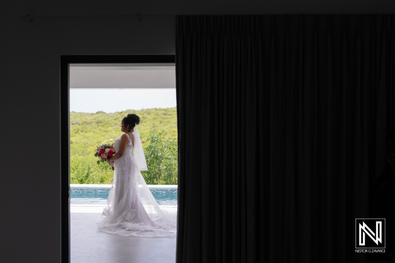 A Bride Holding a Colorful Bouquet Stands in a Doorway Overlooking Lush Greenery and a Serene Pool, Capturing a Moment of Anticipation on Her Wedding Day