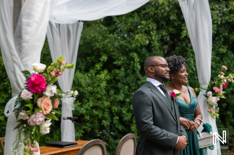 A Groom Stands Proudly Next to His Mother in Elegant Attire During an Outdoor Wedding Ceremony Surrounded by Lush Greenery in the Afternoon Sunlight