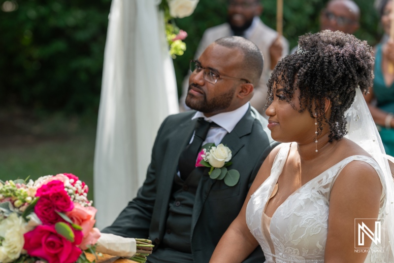 A Couple Listens Intently During Their Wedding Ceremony in a Lush Outdoor Setting, Surrounded by Flowers and Close Friends, Creating a Heartfelt Atmosphere of Love and Commitment