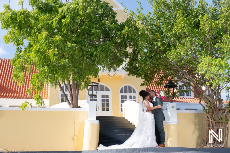 A Couple Shares a Romantic Kiss Outside a Charming Yellow Building Surrounded by Lush Greenery on Their Wedding Day in a Picturesque Location