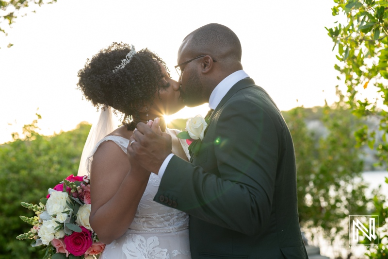 A Joyful Couple Shares a Romantic Kiss During Their Wedding Ceremony at Sunset, Surrounded by Nature and Vibrant Floral Arrangements