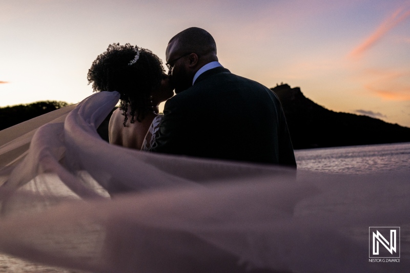 Newlyweds Share a Romantic Kiss at Sunset, With Flowing Veil in the Wind Near a Serene Body of Water Surrounded by Lush Greenery