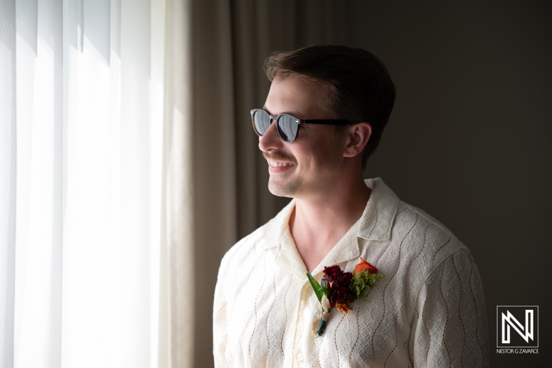 Groom stands by window at Avila Beach Hotel in Curacao before wedding ceremony with family and friends