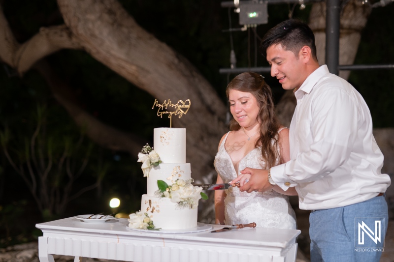 Couple cuts wedding cake at Dreams Curacao Resort during evening ceremony under trees