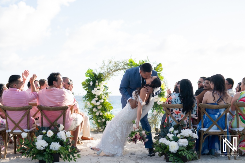 Couple shares a romantic kiss during their wedding ceremony at Playa Hunku in Curacao under a bright sky surrounded by guests