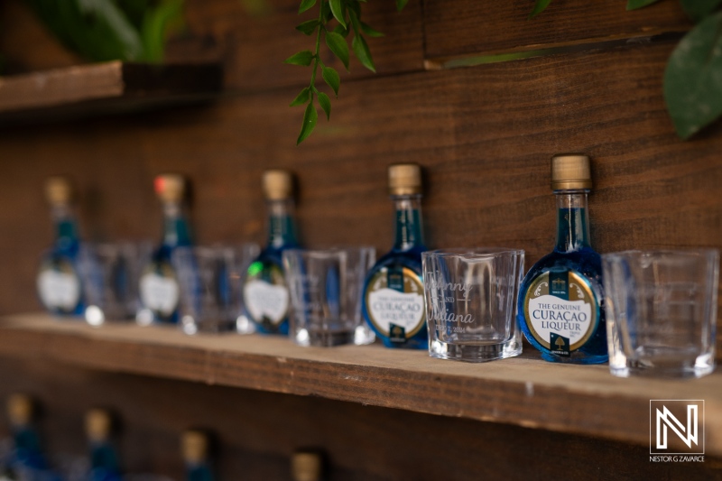 Colorful bottles of Curacao liqueur and shot glasses arranged on a wooden shelf at a wedding celebration by Playa Hunku