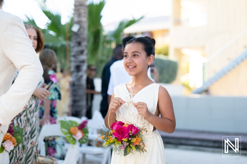 A Young Girl With a Bouquet Smiles Joyfully During a Wedding Celebration at a Beautiful Outdoor Venue in the Afternoon Sunlight