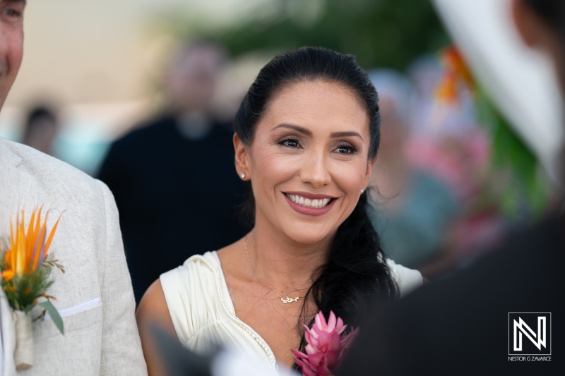 A Joyful Bride Smiles as She Stands With Her Groom During a Wedding Ceremony in a Tropical Garden Setting, Surrounded by Guests and Colorful Decorations in the Warm Afternoon Light