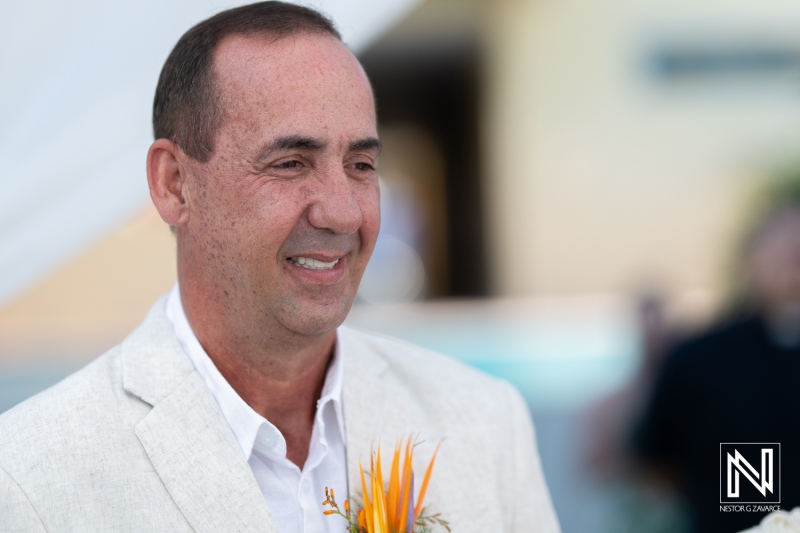 A Groom Smiles Warmly, Dressed in a Light Suit With a Floral Pin, During a Beach Wedding Ceremony Surrounded by Beautiful Decorations and Guests in the Background