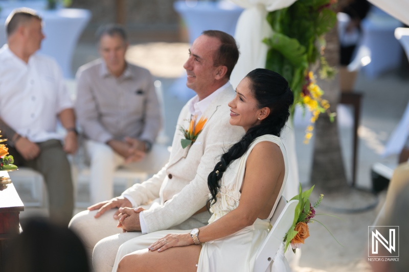 A Couple in Elegant Attire Smiles During Their Outdoor Wedding Ceremony, Surrounded by Guests in a Tropical Setting Adorned With Vibrant Flowers