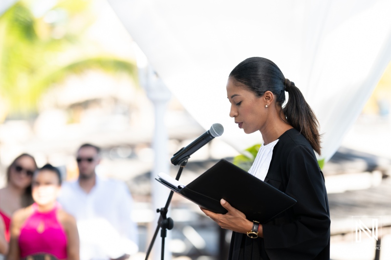 A Woman in Formal Attire Officiating a Wedding Ceremony on a Beach While Guests Attentively Listen in the Background