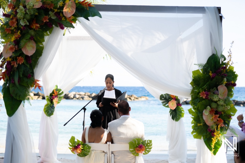 A Serene Beach Wedding Ceremony Featuring an Officiant, a Couple, and Lush Floral Decorations Against the Backdrop of Azure Ocean Waters