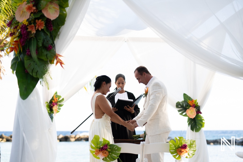 A Beachfront Wedding Ceremony Takes Place Under a Draped Canopy With Floral Decorations as the Couple Exchanges Vows at Sunset in a Picturesque Tropical Location