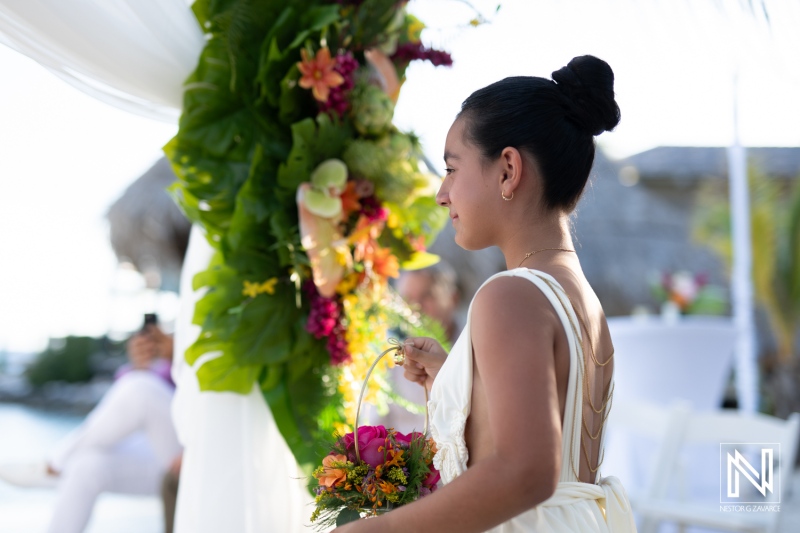 A Young Flower Girl Walks Gracefully Towards the Altar, Carrying a Floral Basket at a Beachside Wedding During Sunset in a Tropical Location