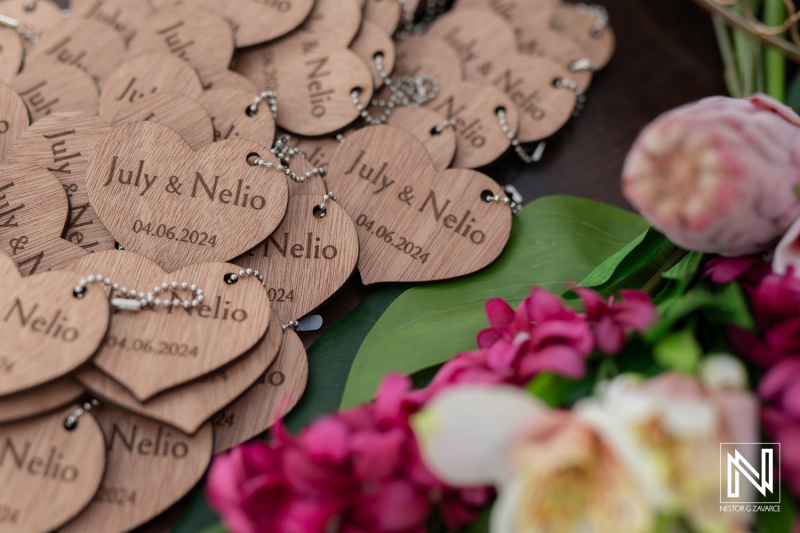 Personalized Wooden Heart-Shaped Tokens for a Wedding Celebration Featuring the Names July and Nelio, Beautifully Arranged With Flowers and a Joyful Ambiance