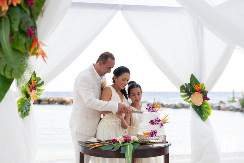 Couple Celebrates Intimate Wedding Ceremony on the Beach, Surrounded by Tropical Flowers and a Beautiful Cake, With Their Child by Their Side During a Sunset