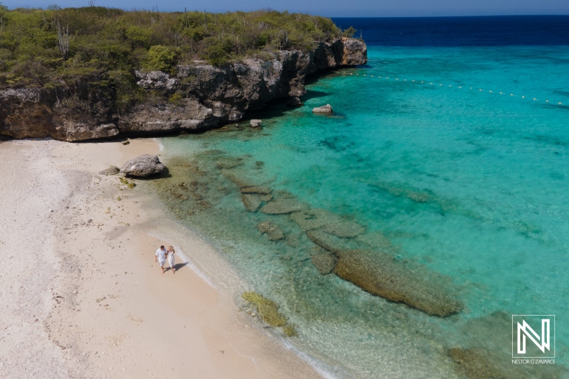 A Couple Walks Hand in Hand Along a Secluded Beach With Clear Turquoise Waters and Rocky Formations, Surrounded by Lush Greenery on a Sunny Day