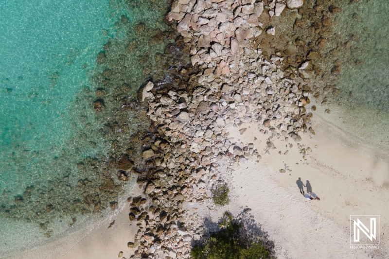 Aerial View of a Rocky Shoreline Meeting Clear Turquoise Waters at a Serene Beach, Capturing Two People Walking Along the Sandy Coast During a Sunny Afternoon