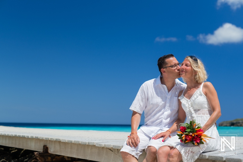 A Joyful Couple Shares a Tender Kiss on a Sunny Beach Pier While Holding a Bouquet of Tropical Flowers Under a Bright Blue Sky