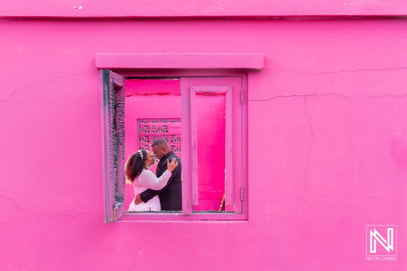 Couple shares a romantic kiss framed by a vibrant pink window in Curacao during their wedding celebration