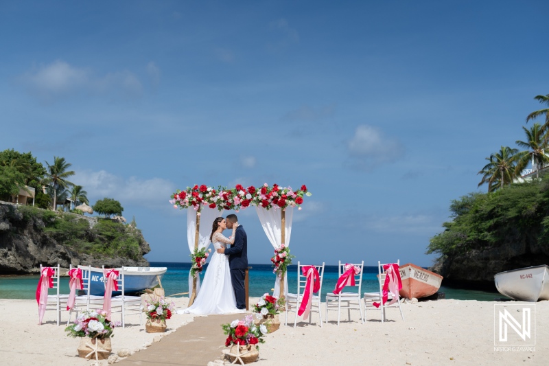 A Couple Exchanges Vows Beneath a Floral Arch on a Picturesque Beach With Colorful Boats in the Background During a Bright, Sunny Day