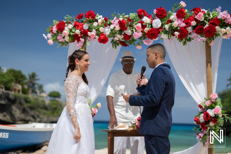 A Couple Exchanges Vows Under a Floral Arch on a Beach During a Sunny Wedding Ceremony, Surrounded by Lush Greenery and a Calm Sea in the Background