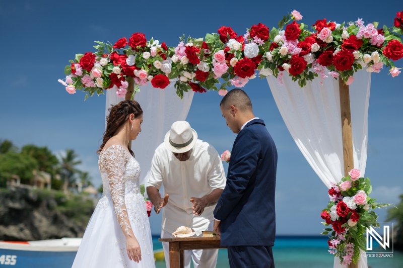 Couple Exchanging Vows Under a Floral Arch on a Beach During a Sunny Day, With an Officiant Standing Before Them in a Tropical Setting