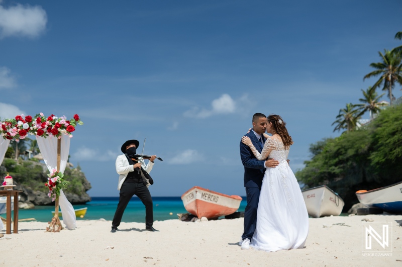 A Couple Shares a Romantic Moment During Their Beach Wedding Ceremony With a Violinist Performing in the Background on a Sunny Day at a Tropical Location