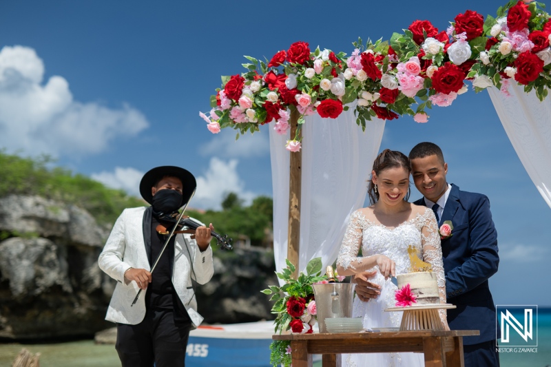 A Joyful Beach Wedding Celebration With a Couple by a Decorated Arch, a Musician Playing Violin, and a Beautiful Cake Under a Clear Blue Sky