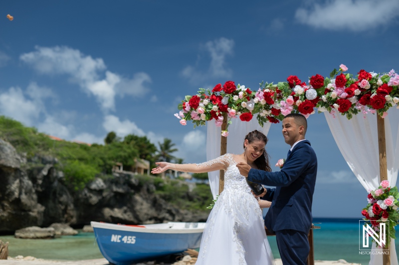 A Joyful Couple Celebrating Their Wedding on a Picturesque Beach With Floral Decorations and a Boat in the Background Under a Bright Blue Sky