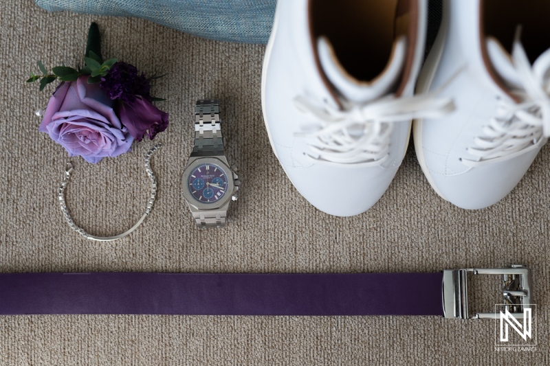 Stylish accessories layout featuring white sneakers, a silver watch, a purple belt, a bracelet, and a rose on a textured surface