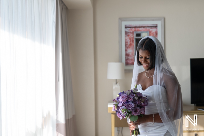 A bride in a beautiful white gown smiles while holding a bouquet of purple flowers in a bright room before the wedding ceremony