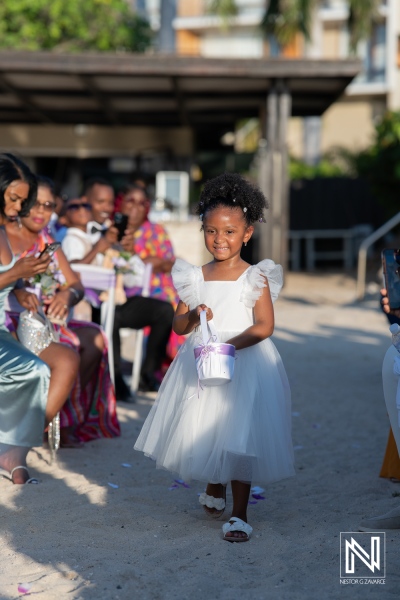 A young girl in a white dress walks down the sandy aisle during a beach wedding ceremony in the late afternoon, surrounded by guests joyfully taking photos