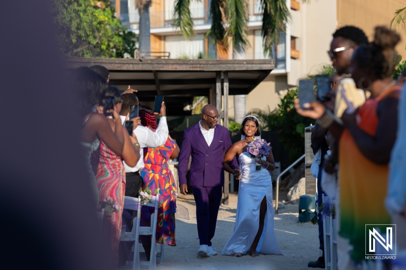 A beautiful bride in a silver gown walking down the aisle with her father during a summer outdoor wedding ceremony in a tropical garden setting