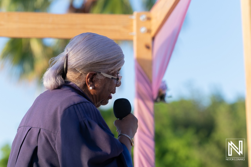 Elderly woman speaking at a community event outdoors, addressing the audience with enthusiasm during daytime beneath a clear sky surrounded by greenery