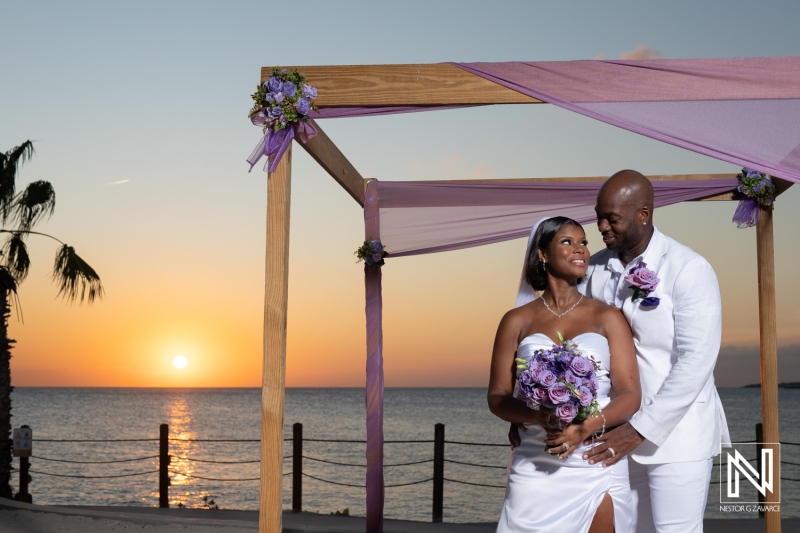 A couple celebrates their wedding at sunset by the beach with vibrant purple flowers in a serene coastal setting