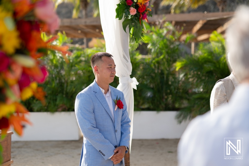 Ceremony at Kokomo Beach in Curacao features a groom awaiting his bride beneath vibrant floral decorations