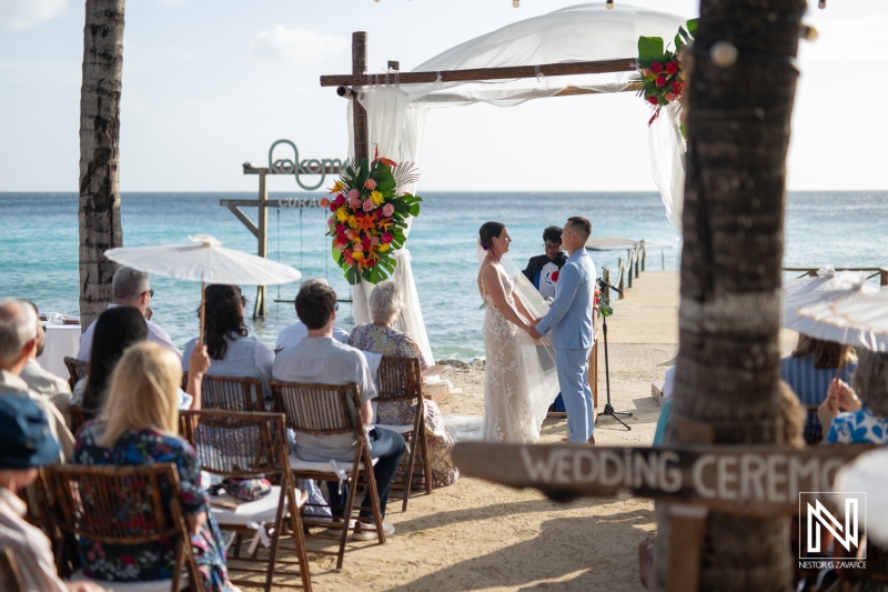 Wedding ceremony takes place at Kokomo Beach in Curacao with ocean backdrop and guests enjoying the beautiful setting under a clear sky