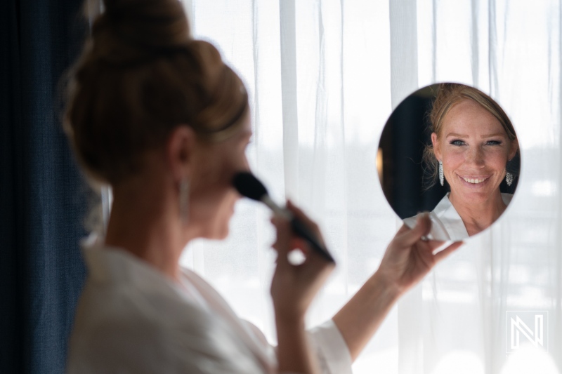 A woman applies makeup while looking into a mirror, preparing for a special event near a window in soft natural light during the day