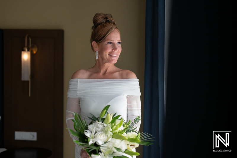 A radiant bride stands by the window holding a lush bouquet in an elegant white gown, capturing a moment of joy before her wedding ceremony in a stylish indoor setting