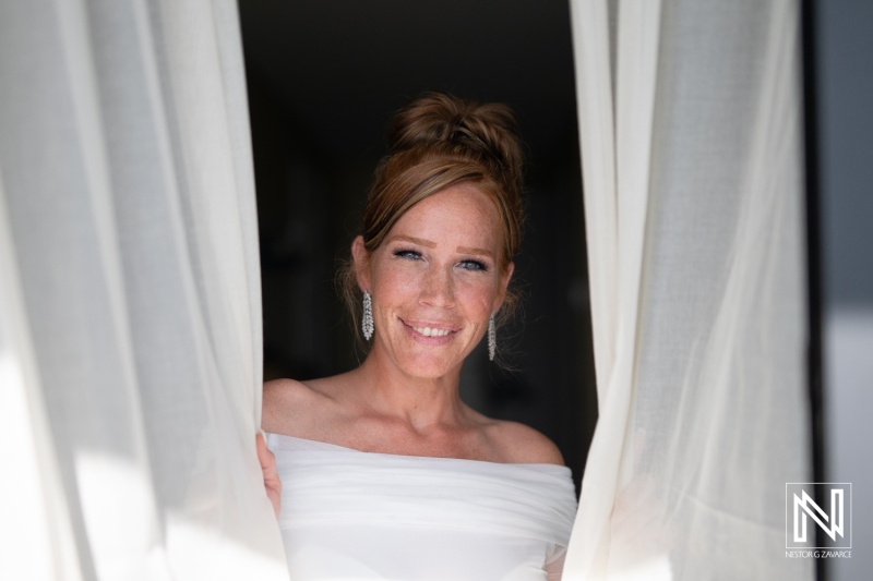 A smiling bride wearing an off-shoulder wedding dress stands by the window, capturing the joy of her special day in a bright, sunlit room