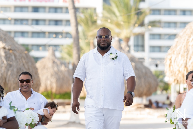 A groom walks confidently towards the beach altar at a tropical wedding ceremony with guests dressed in white during a sunny afternoon