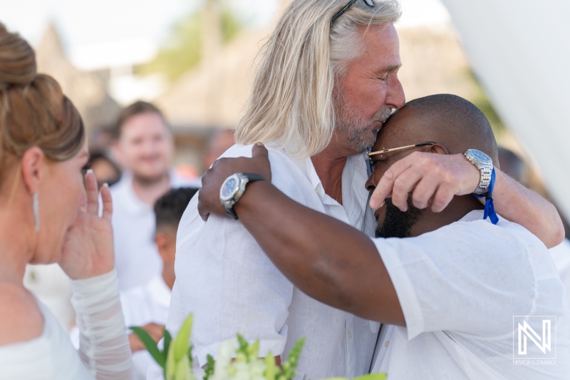 Two close friends embrace warmly during an outdoor wedding ceremony under a bright sky in a tropical setting surrounded by guests dressed in all white