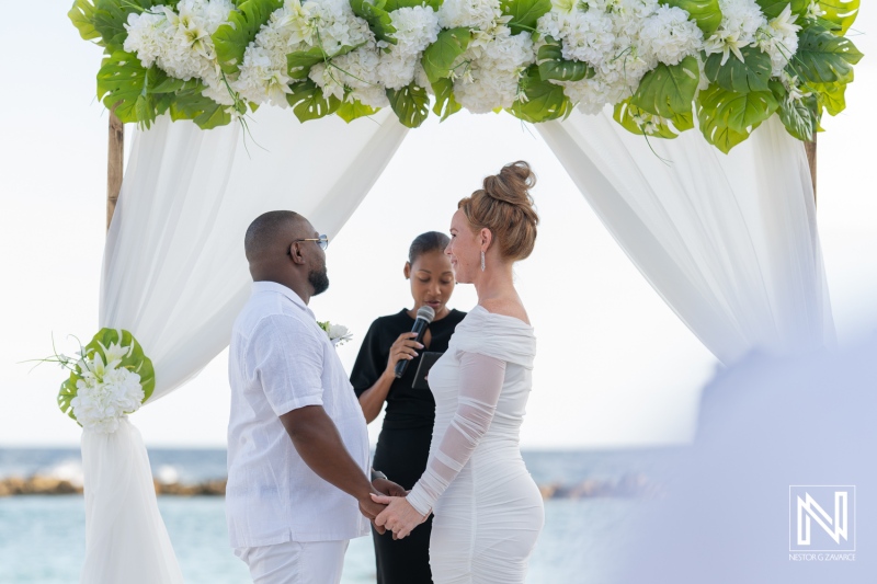 A couple exchanging vows under a beautiful floral arch during their beach wedding ceremony in the Caribbean at sunset, surrounded by nature and love