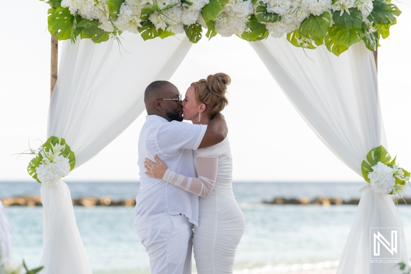 A joyful couple exchanges vows at a beachside wedding ceremony under a floral arch on a sunny day with a beautiful ocean backdrop