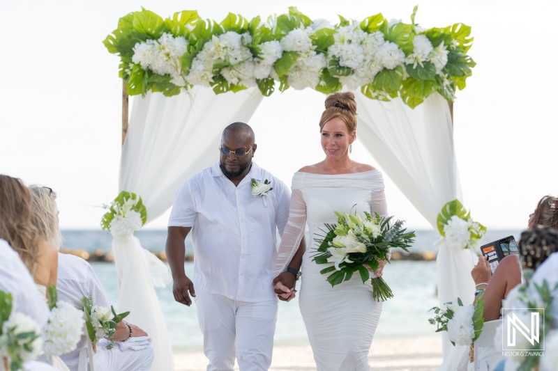 A couple walks hand in hand under a floral arch on the beach during their wedding ceremony against a stunning ocean backdrop on a sunny day