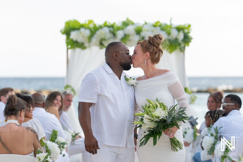 Romantic beach wedding ceremony at sunset featuring a couple sharing a kiss while surrounded by guests and floral arrangements in the Caribbean